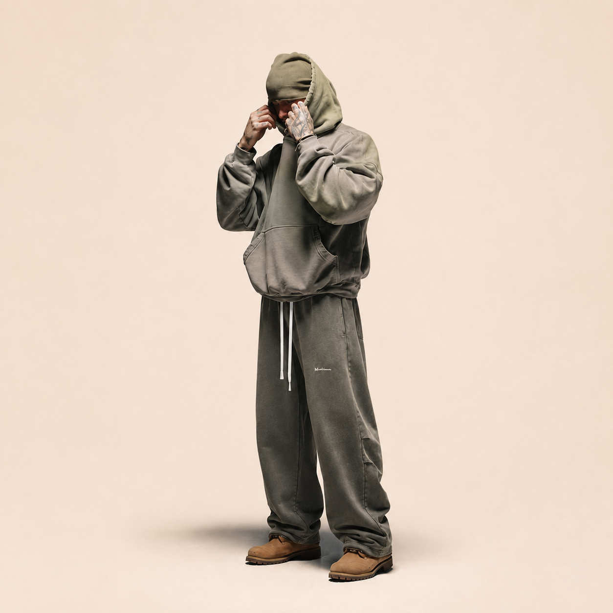A person modeling an oversized matching hoodie and sweatpants set in a washed olive green and dark gray colorway, standing against a solid beige background.